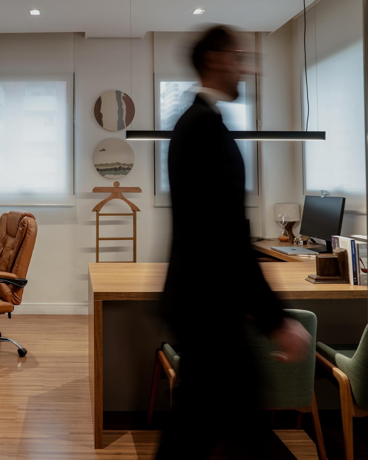 Advogado de terno escuro atravessa, em movimento, uma sala do escritório com mesa de madeira e janelas claras.