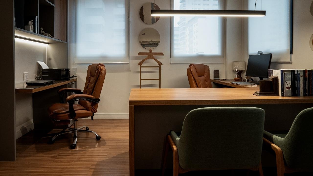 Sala de atendimento do escritório, com mesa de madeira, poltrona de couro e cadeiras verdes.