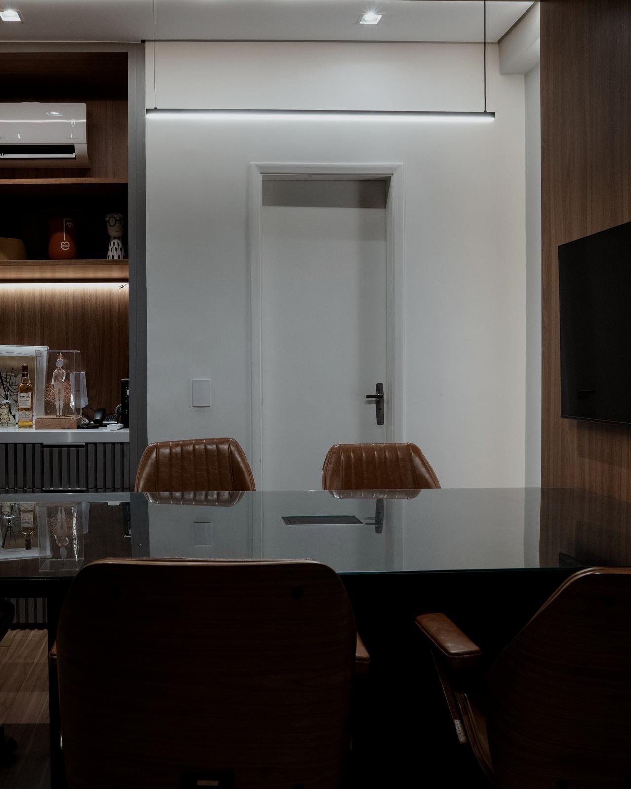 Sala de reunião do escritório, com cadeiras de couro caramelo em torno de uma mesa de vidro escuro e armário de madeira iluminado.