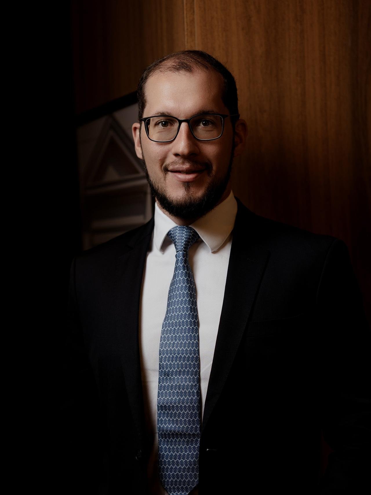 Guilherme Sivieri Reyna, de óculos e barba, terno escuro e gravata azul, diante de uma parede de madeira.