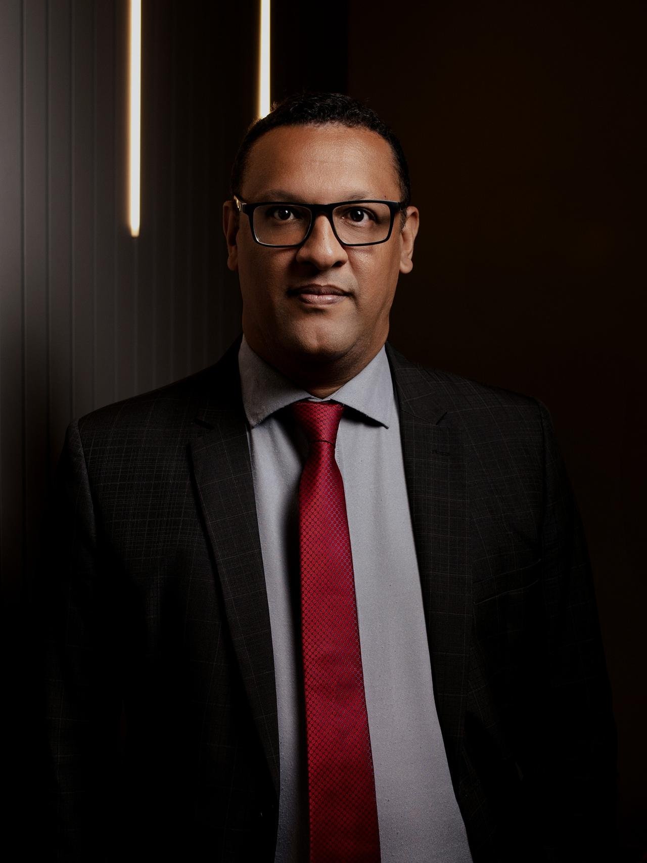 Paulo Henrique dos Santos, de óculos, terno grafite e gravata vermelha, diante de um painel ripado com luz vertical.
