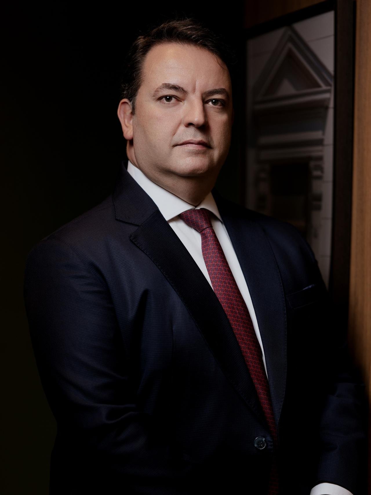 Ricardo Calil Haddad Atala, de terno azul-marinho e gravata bordô, em frente a uma parede de madeira.